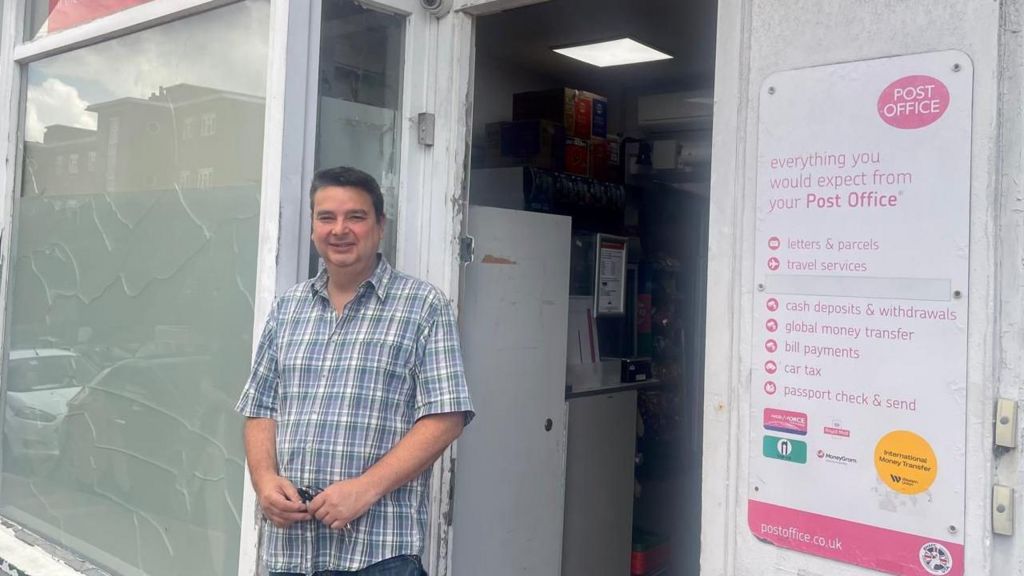 A man with dark hair and a check shirt stood outside of a white shop. The man is smiling.