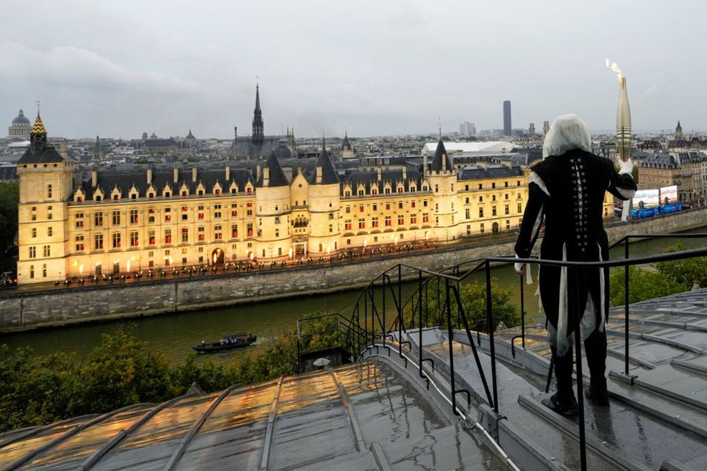 A hooded and masked torchbearer carries the Olympic flame over a building along the Seine River in Paris, France, during the opening ceremony for the Paris 2024 Olympic Games.