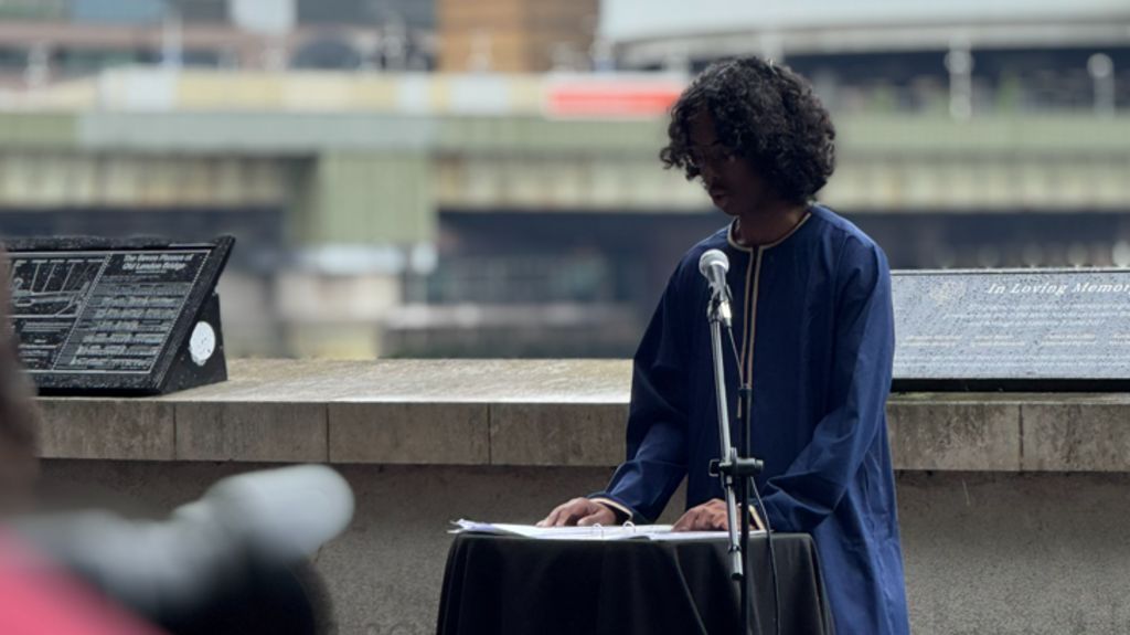 A person speaks at a microphone beside a small table and  a memorial plaque and riverfront setting appear behind them.