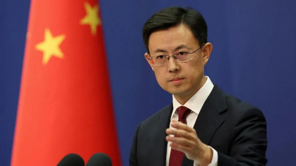 An image of Chinese Foreign Ministry spokesperson Guo Jiakun taking a question from a journalist at a press conference in Beijing, China. He is wearing a black suit jacket with a white button-up shirt and a deep red tie. He has short, black hair that is parted on the right, and is wearing frame-less eye-glasses. He is standing at a podium. To his left is a blurred out flag of China.