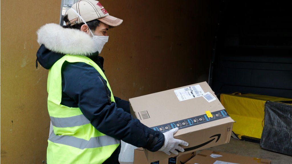 A delivery man wearing a protective mask carries an Amazon package.