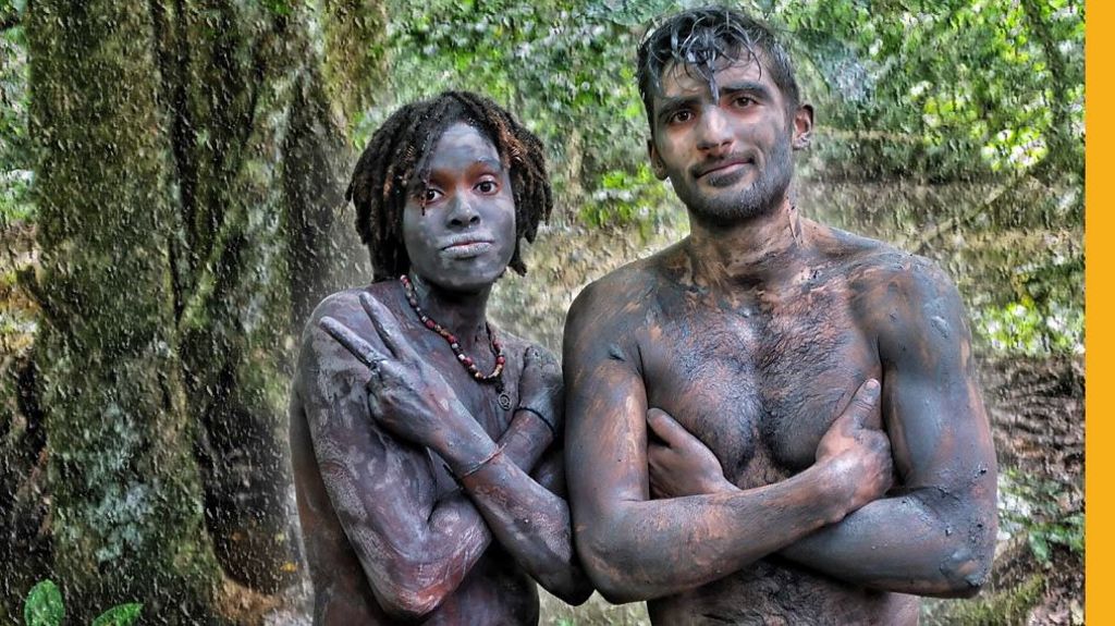 A woman and Ben Zand stand in the jungle topless, posing with arms folded, and with blue clay on their skin.