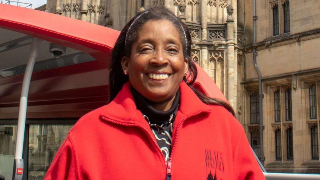 Pamela Roberts smiles at the camera, she's wearing a red zip up fleece which has a Black Oxford logo on it. She is standing on a bus with a historic Oxford building in the background