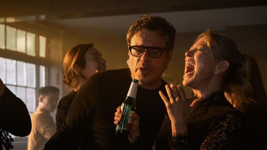 Jemaine Clement and Nicola Walker as Steve and Alice singing holding a beer bottle 