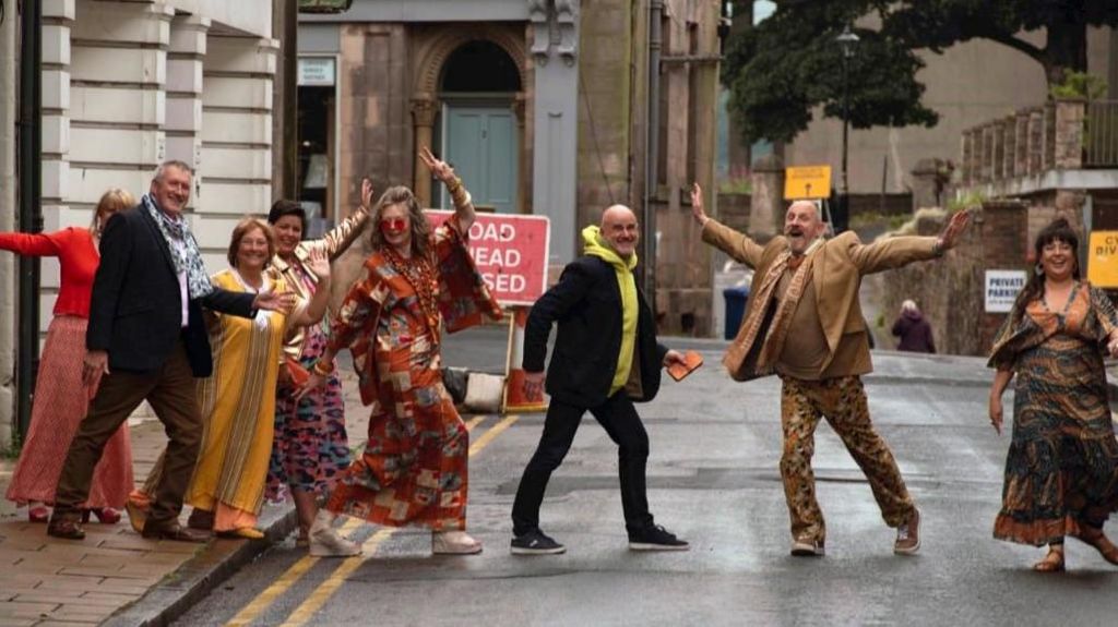 A group of people all wearing bright and unusual clothes walking across the road.