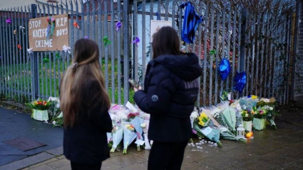 Photo of two girls from behind, stood next to flowers near to the scene in south Bristol where two teenage boys, aged 15 and 16, died after a stabbing attack.
