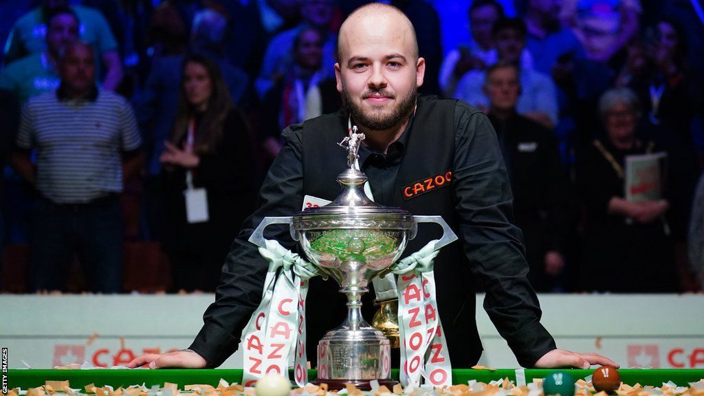 Luca Brecel is looking at the camera dressed in a black shirt smiling with his hands resting on the snooker table. The silver trophy with the sponsors red and white tassles is directly in front of him.