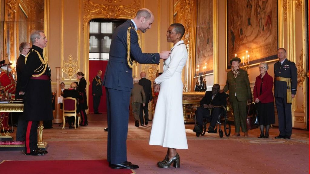 A man in a navy suit presenting an award to a lady in a white dress. They are at a ceremony inside a castle.