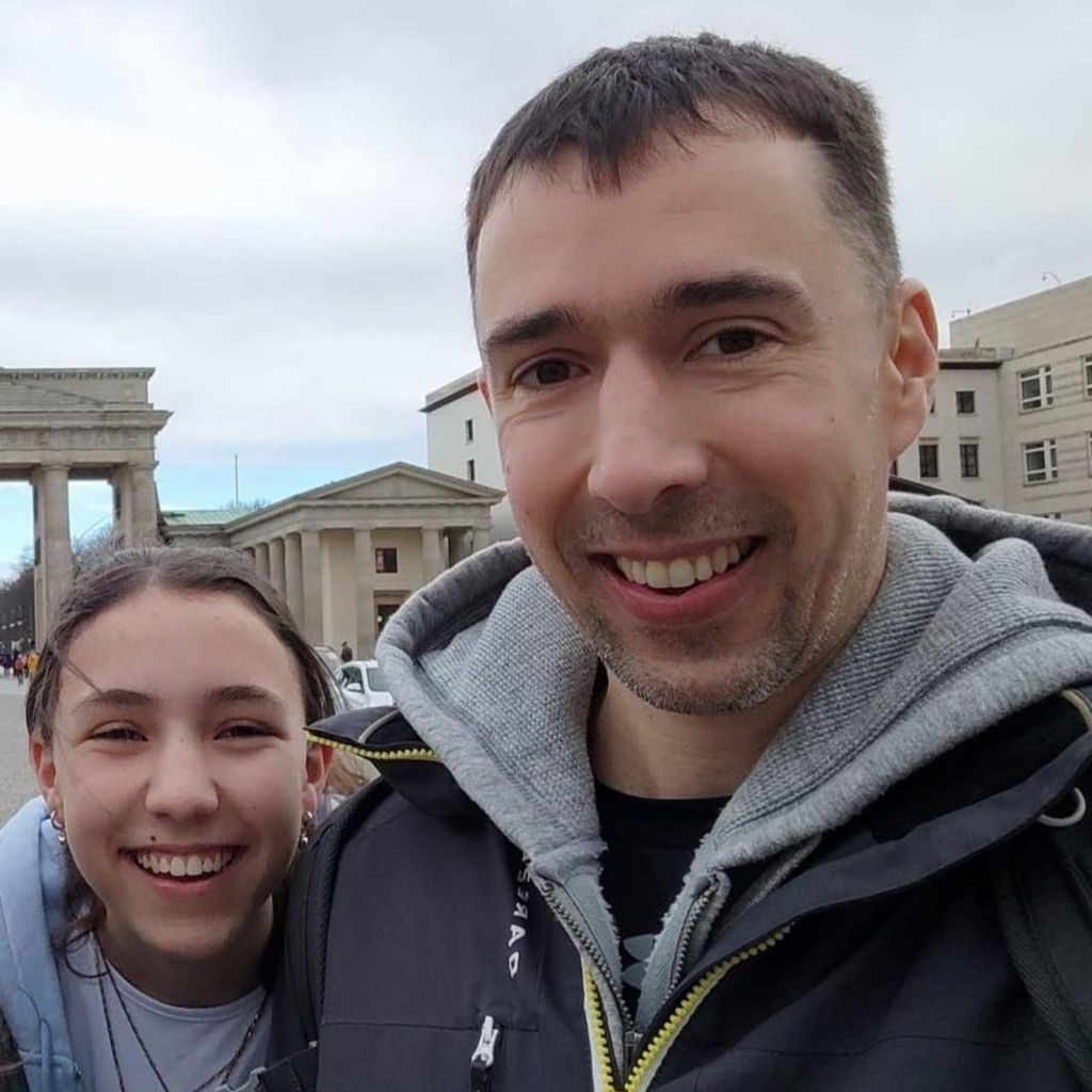 Sqn Ldr Mark Long and his daughter, who are both smiling and wearing hoodies, stand in front of classical buildings in Berlin.