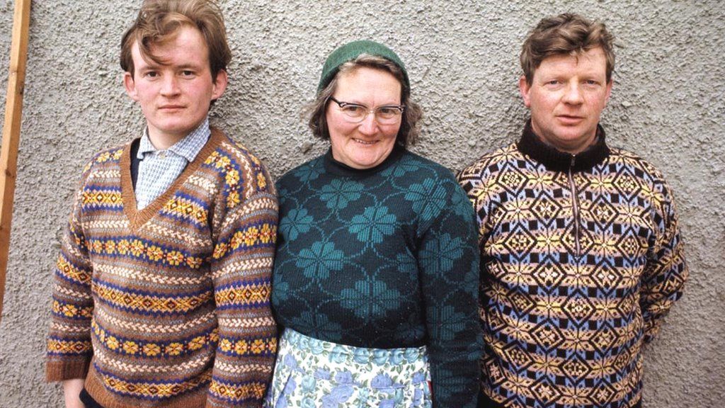 A woman and two men pose wearing Fair Isle jumpers in front of the wall of a cottage on one of the Shetland Islands in 1970.