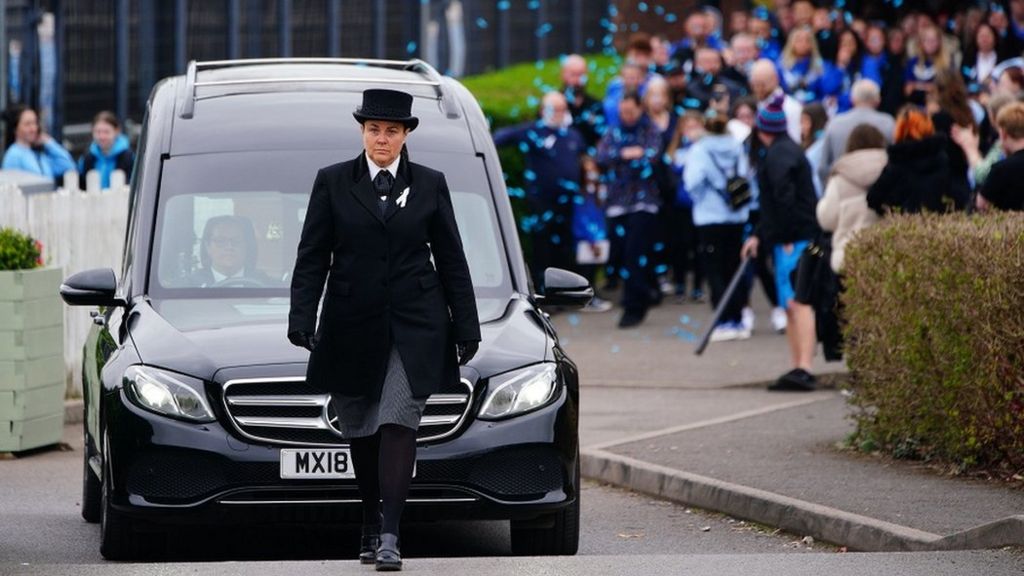 An undertaker walks in front of a hearse while a crowd stands in the background
