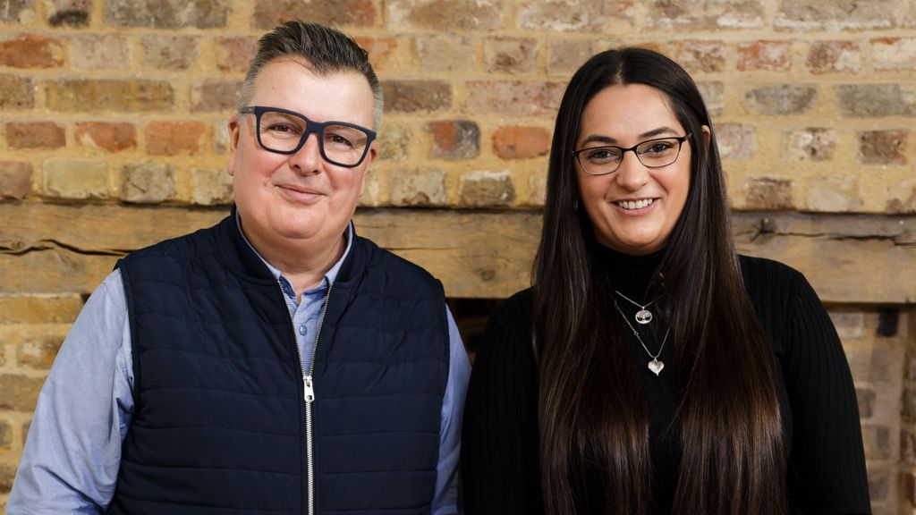 Paul Richards and Karima Motala stand side by side indoors against a brick wall, both facing the camera.