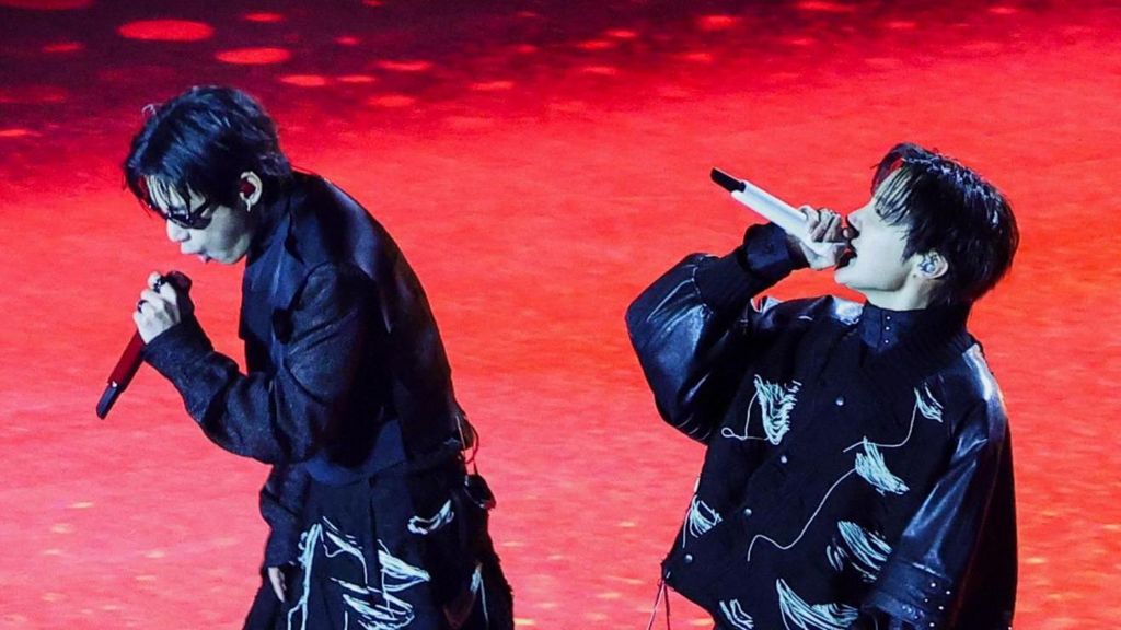 Two members of K-pop group BTS wearing black jackets and holding mics perform on a red stage