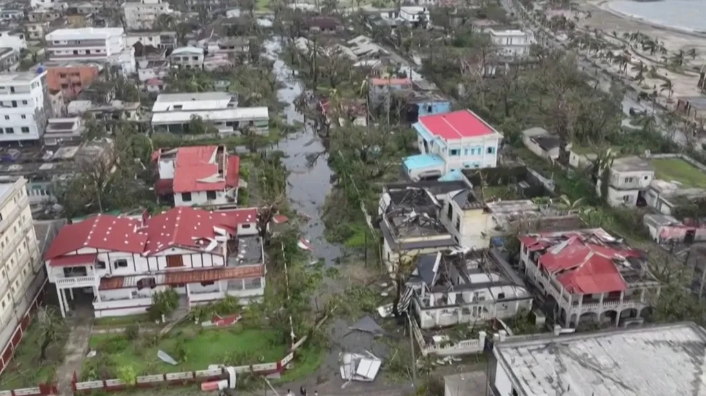 Aerial view of a coastal neighbourhood in Madagascar after a cyclone, showing widespread destruction. Many buildings have damaged roofs, fallen trees block the streets, and floodwater fills the roads. Debris is scattered throughout the area, with the shoreline visible in the background.