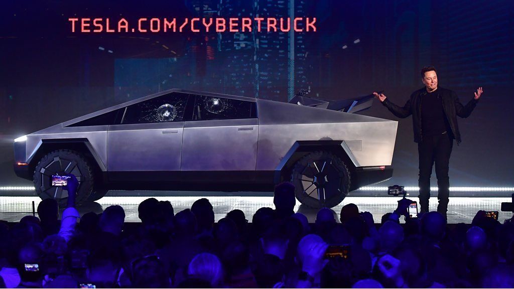Elon Musk shrugs while standing beside a Cybertruck with two broken windows