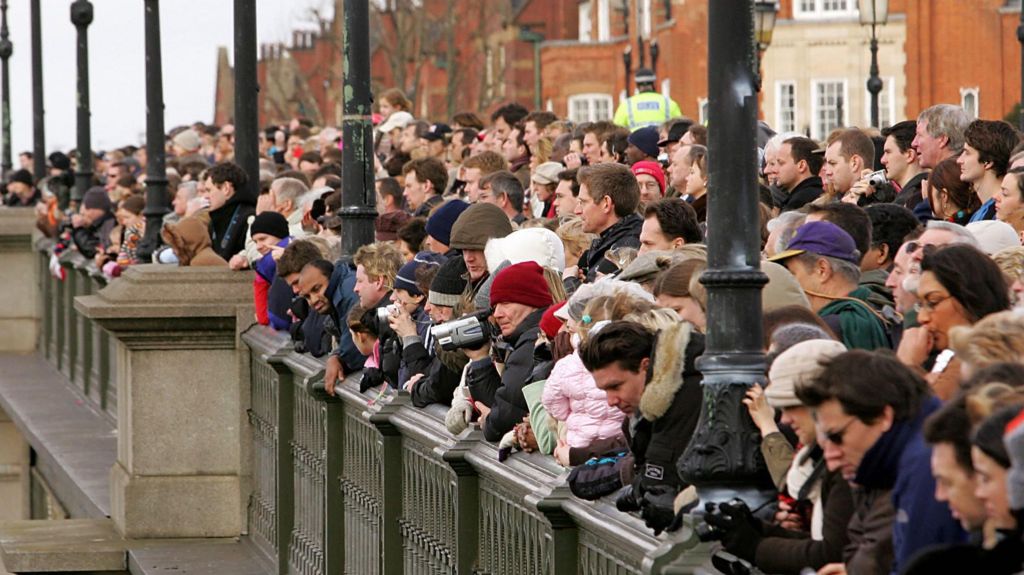 Hundreds of people are packed behind the fence beside the Thames in west London. Many are holdings cameras