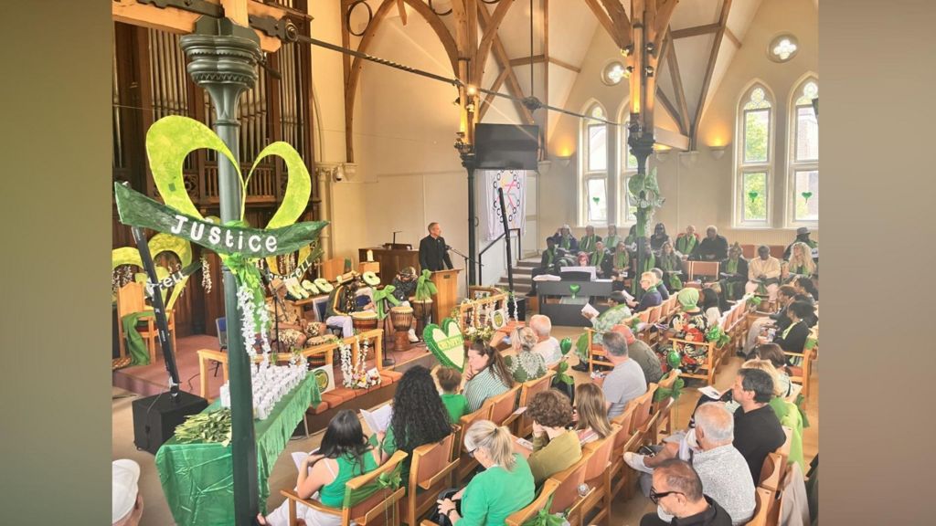 A large congregation sit in a church which is decorated with green banners.