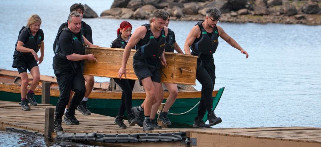 Contestants on the traitors carrying a coffin along a platform