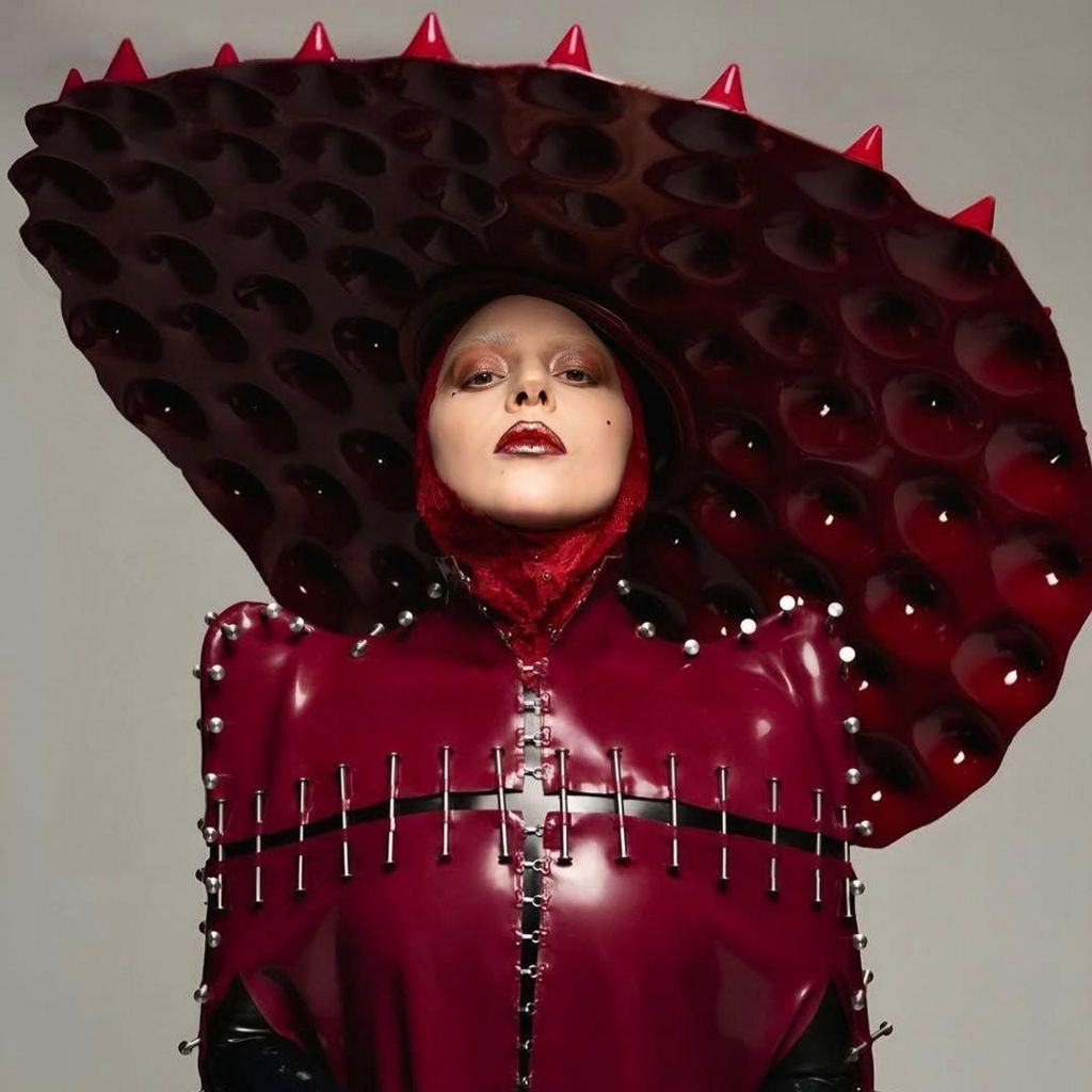 Lady Gaga wears a blood-red plastic leather outfit, stitched togeter with nails, and topped by an oversize disc-shaped hat