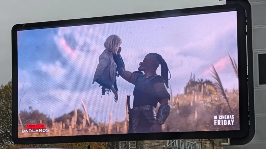 An advert for the film Predator Badlands shows a large, alien-like figure holding up a smaller, human-like figure which has been severed in two and has its decapitated spine showing.