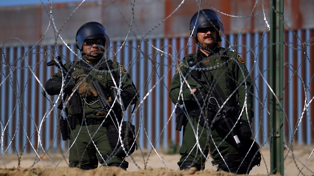Two men with guns and helmets behind barbed wire fence, security patrol at border