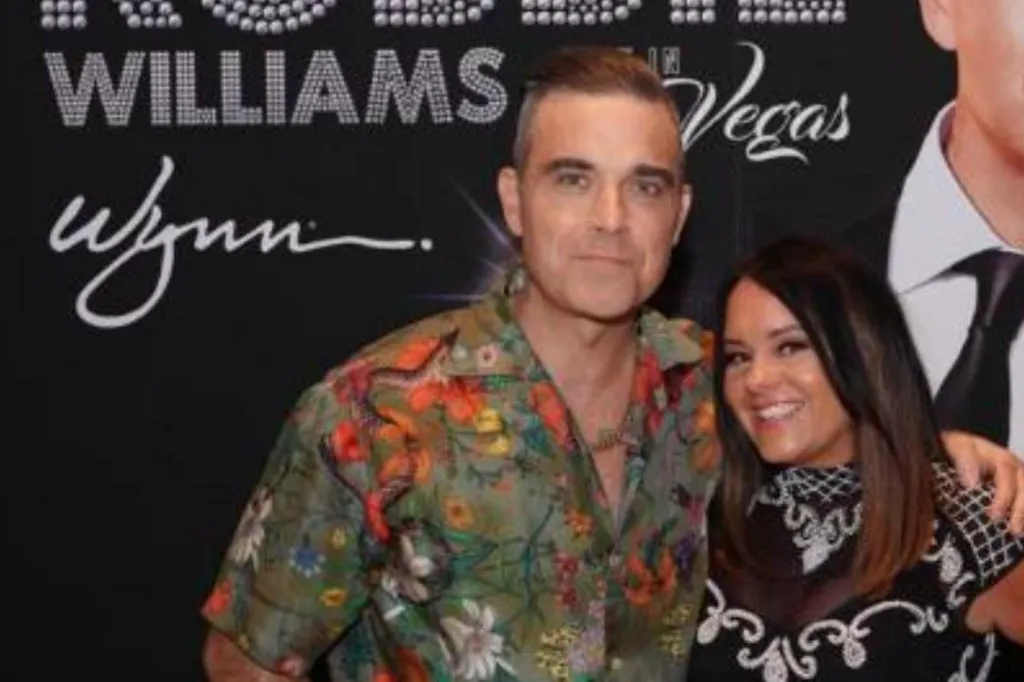 Robbie Williams stands next to a woman and has his arm around her. Williams wears a green tropical shirt and the woman, who has brown hair, wears a black and white dress. They are in front of a poster of Williams.