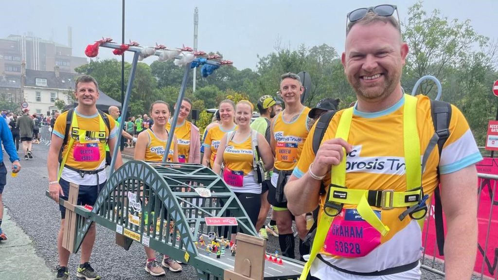 A group of seven runner in yellow T-shirts smiling at the camera. They hold a mini model of the Tyne Bridge, showing the start of the with the Red Arrows flying above it. Derick Jackson is at the back.