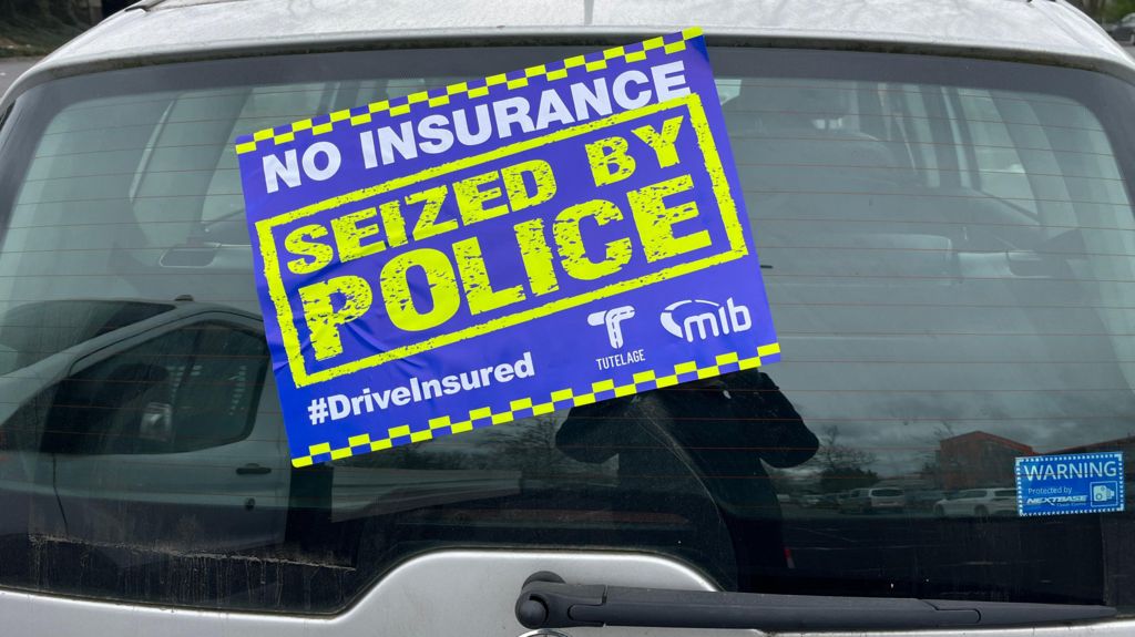 Silver car with a blue yellow and white sticker covering the rear window saying it's been seized by police for having no insurance.