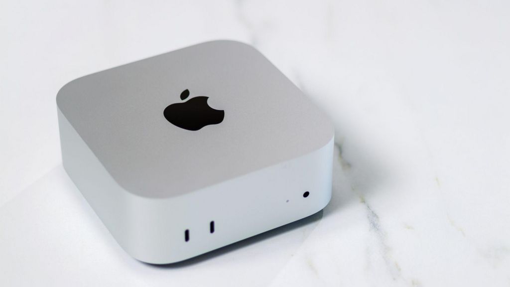 A matte, silver-coloured cube with rounded corners and a black Apple logo sits on a white marble countertop.