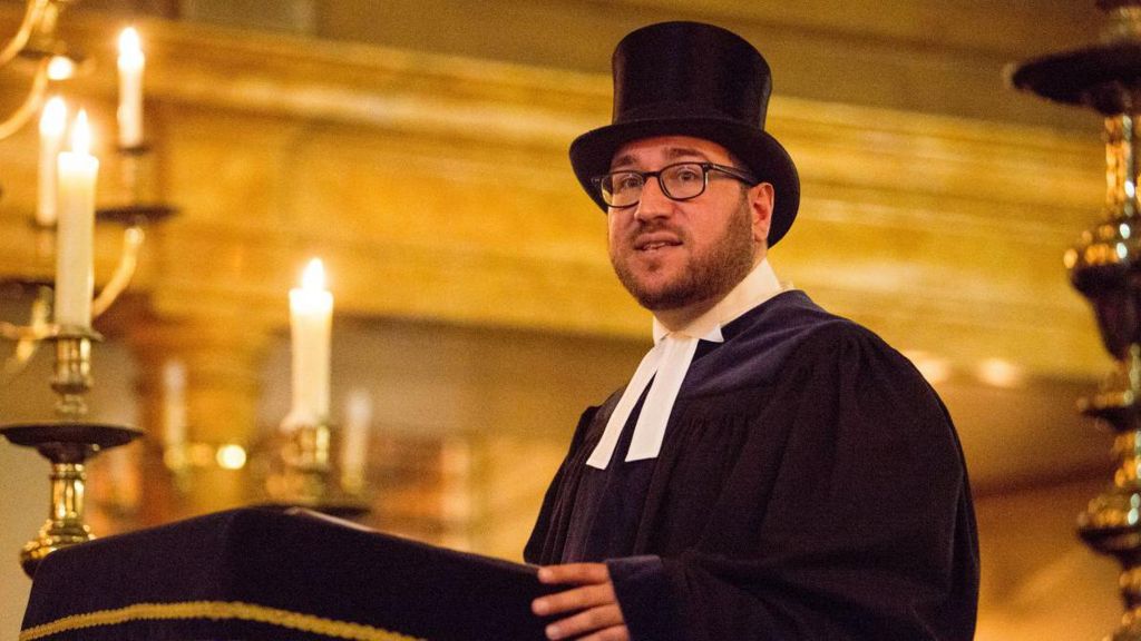 an image of Rabbi Shalom Morris wearing a hat and in robes inside a synagogue