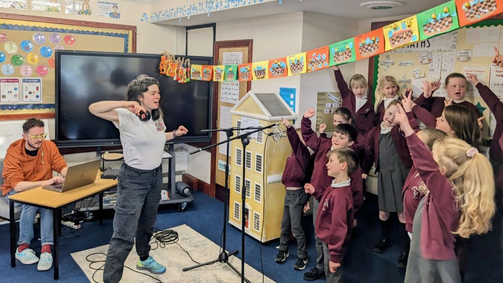 Chloe Shimmin standing in a classroom in front of a group of school children while are holding pointing a finger up into the air. She has dark brown hair and is wearing a light grey T-shirt, dark grey trousers and blue trainers and has a pair of headphone around her neck. The children are wearing maroon and grey school uniforms and are standing in front of two microphones on stands. And man wearing an orange hoodie and blue jeans sits at a table in the background looking at a laptop, with a large black TV screen on a stand next to him. The room has bright boards with bright note on them, colourful paper bunting hangs above the children.