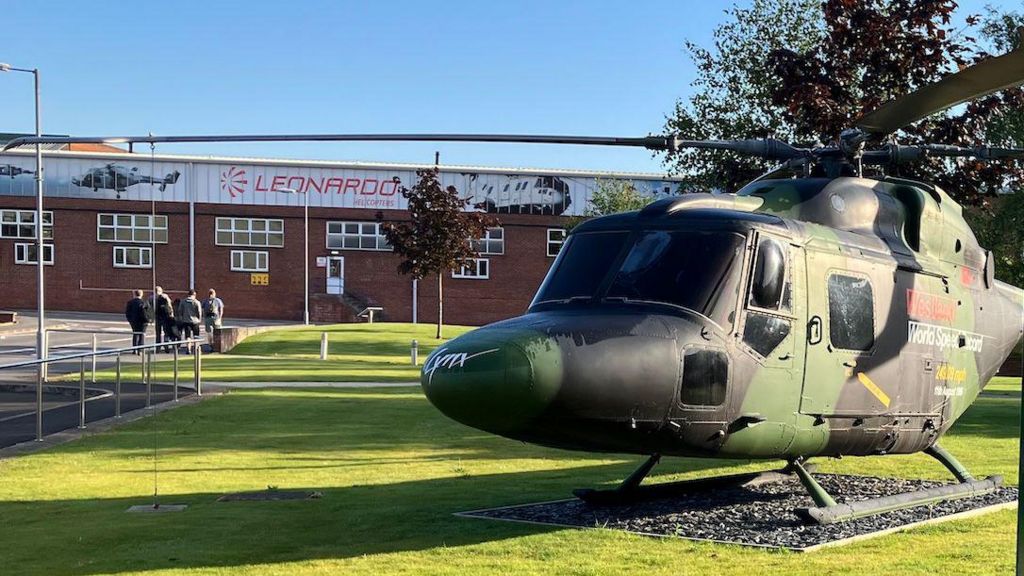 A camo-painted helicopter standing on a green field with a factory building in the background. The word Leonardo is painted across the top of the building.