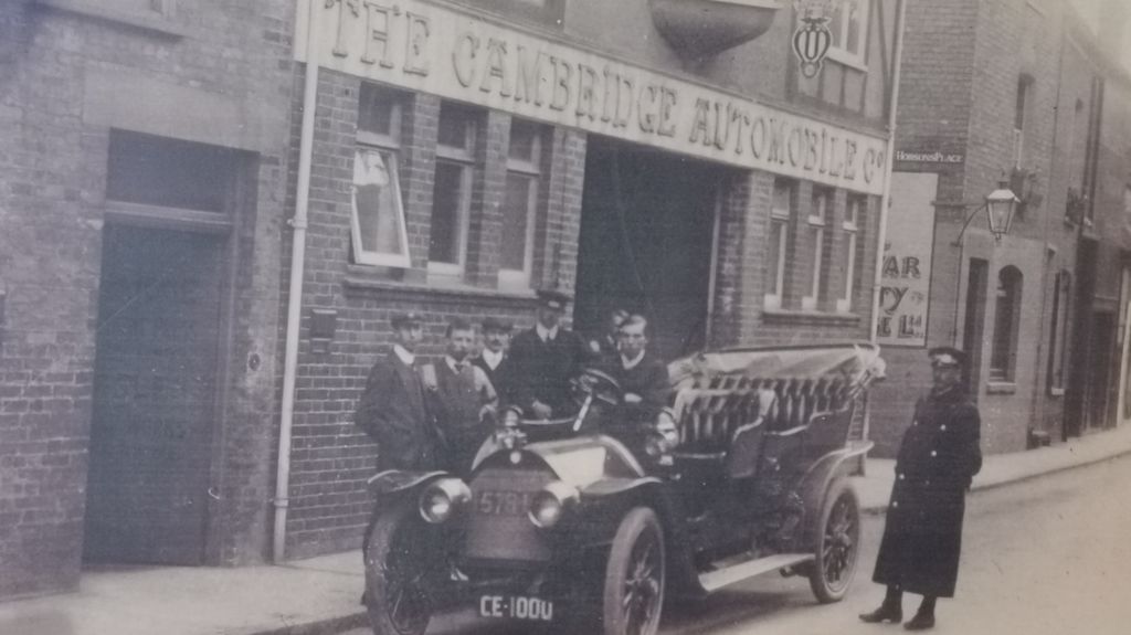 7 men standing by a 1900s car outside a large building with The Cambridge Automobile Company written on the sign. Two of the men wearing police uniforms