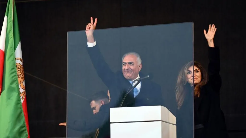 Reza and Pahlavi and his wife wave to crowds on stage. They appear to be standing behind a glass screen. A red, green and white flag can be seen on the left.