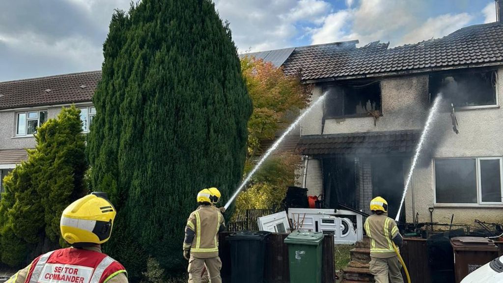 Fire crews with two water hoses putting out a terraced house fire that has severely burnt rendering. The white front door has been removed and placed on its side. There is a big hole in the roof and black smoke is coming out of the windows.