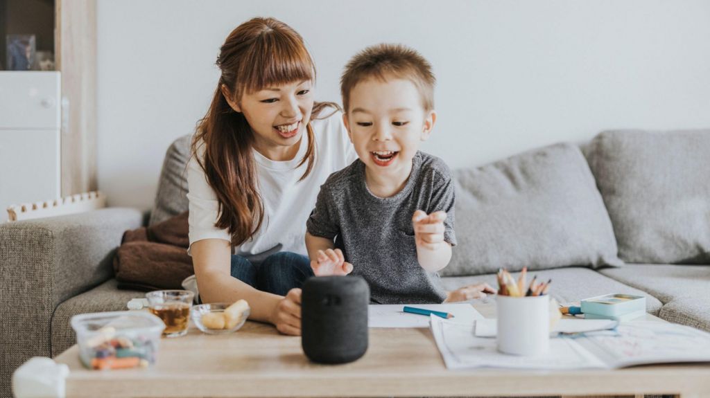 A young mother and her young son speak to a smart speaker on the table in front of them