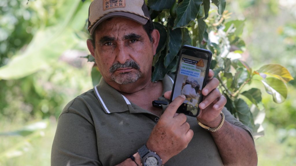A man stands outside holding up a phone displaying a picture of his grandson