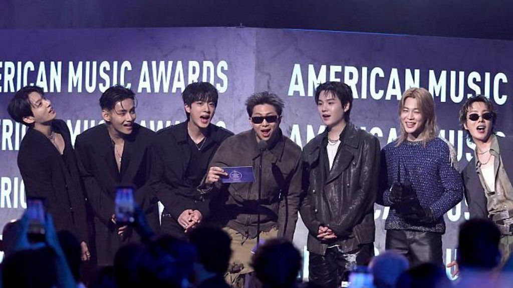 Seven members of BTS stand on stage in front of the words American Music Awards in white text.