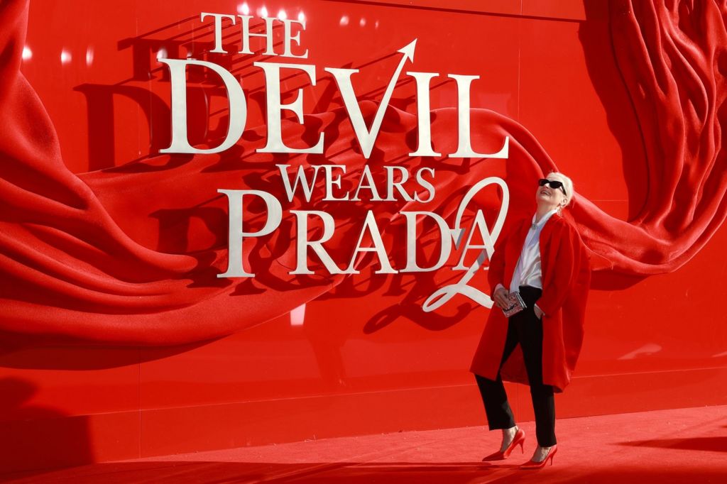 Meryl Streep stands smiling on the red carpet next to a "The Devil Wears Prada 2" sign.