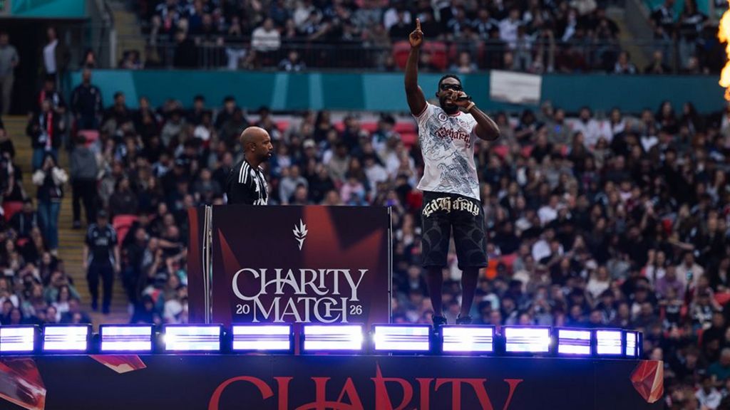 Tinie Tempah on stage on the pitch during the half-time show, jumping up with a microphone in one hand and his other arm raised. Thousands in the crowds are watching him.