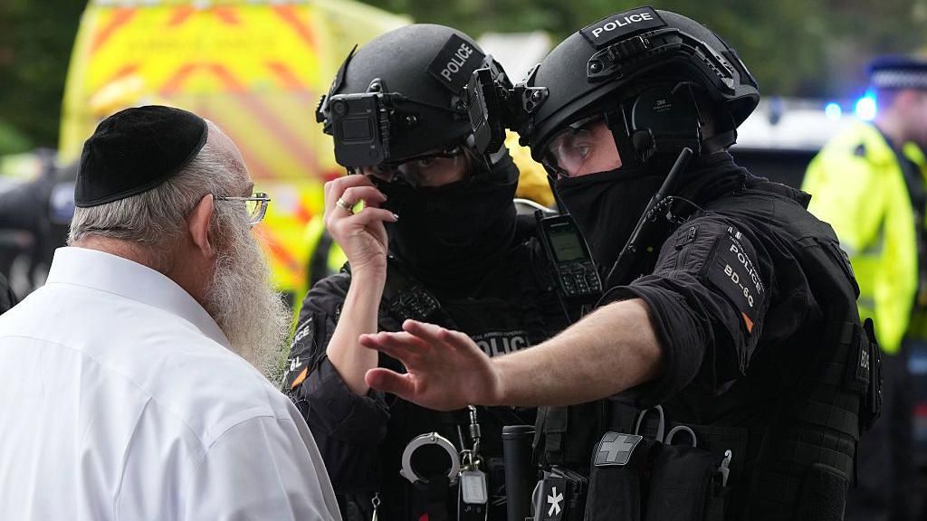 A man with a long white beard, and wearing a white shirt and a cloth kippah, speaks to two police officers, both wearing black uniforms and helmets.