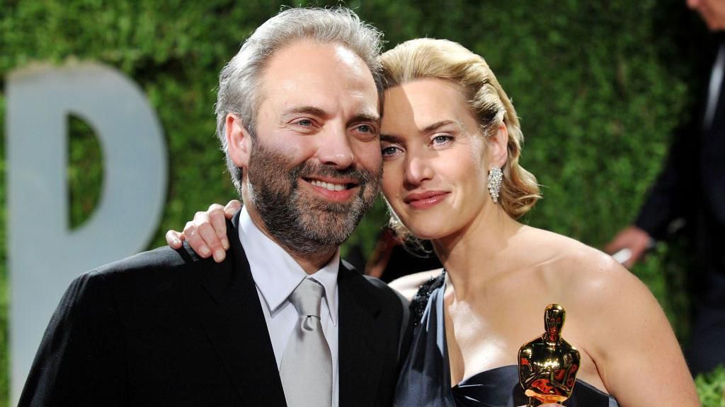 Winslet and Mendes smile while their heads are touching. Winslet holds a golden Oscar statue.
