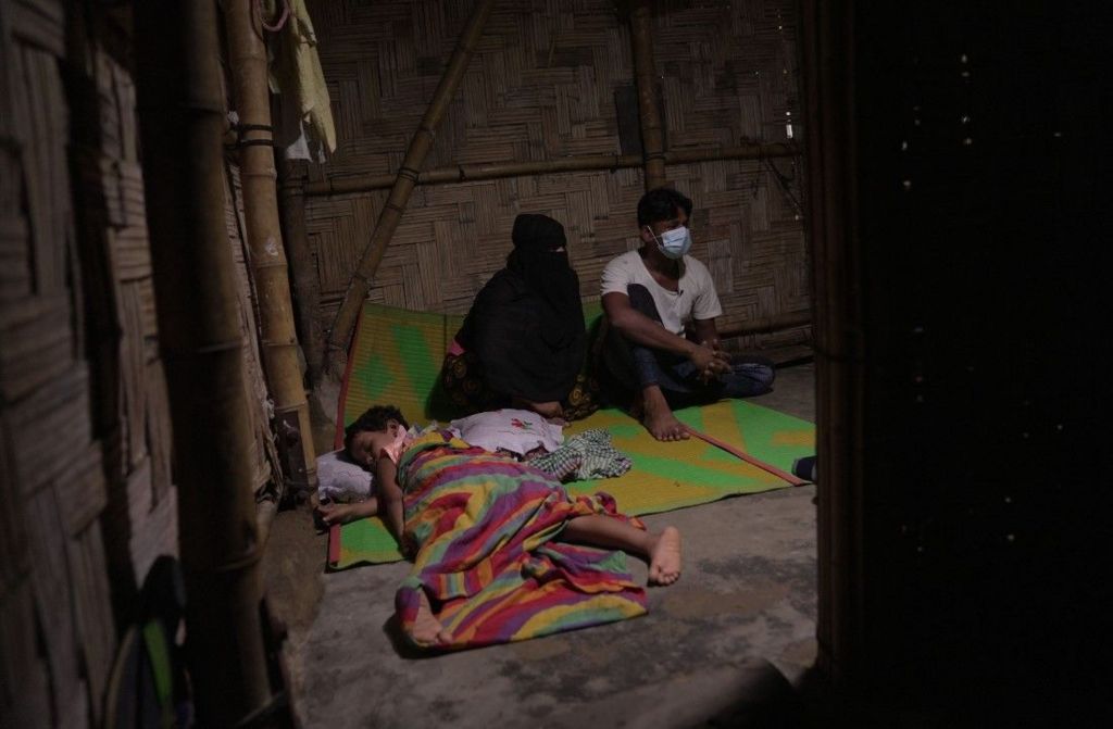 Three people are visible in a hut which appears to be made of bamboo. A child lies on the floor, covered by a colourful blanket, with a man and a woman sitting next to them on a green mat. The woman is wearing a head and face covering, and the ban has a medical face covering on. 