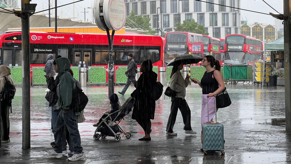 People stand under a shelter in Newham while it rains. There are buses in the background.