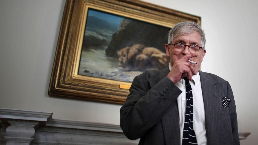 David Hockney smoking in an art gallery in 2011. He is standing in front of an old master painting while doing so.