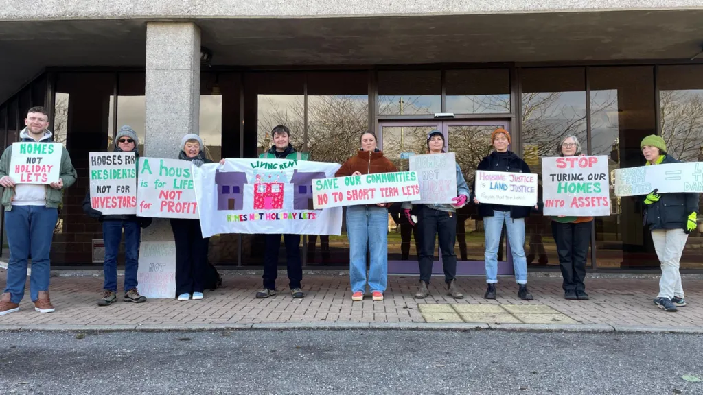 Nine people are standing in a row outside the glass fronted entrance to Highland Council's headquarters in Inverness. The people are holding up signs and banners.
