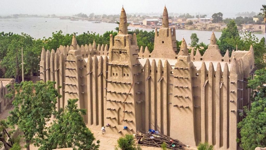 The giant mud structure of the Grand Mosque of Mopti surrounded by trees, with a river seen in the background.