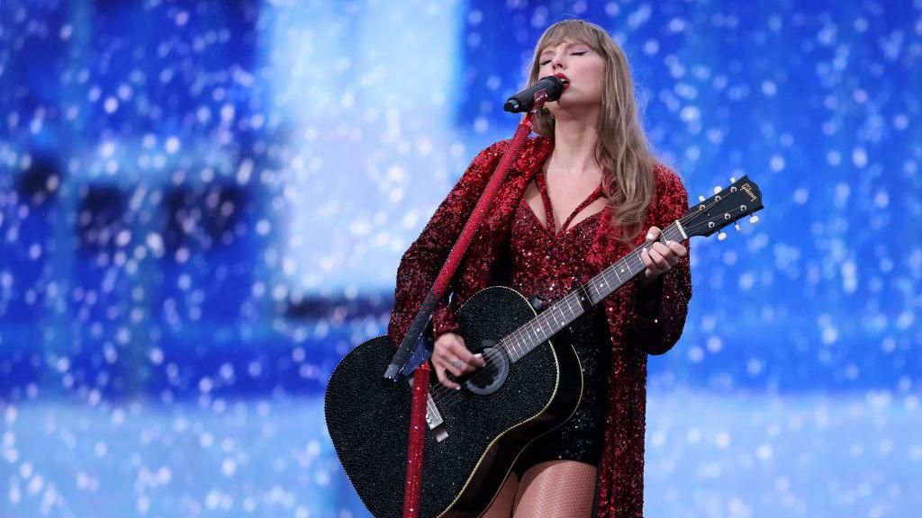 Taylor Swift performing on stage. She is playing a black guitar and singing in to a microphone. She is wearing a red sparkly outfit with a red sparkly jacket.