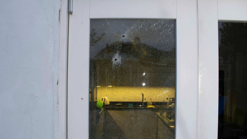 A glass door has two bullet holes and damage to the glass. Through the door a kitchen counter with a kettle and tap can be seen.