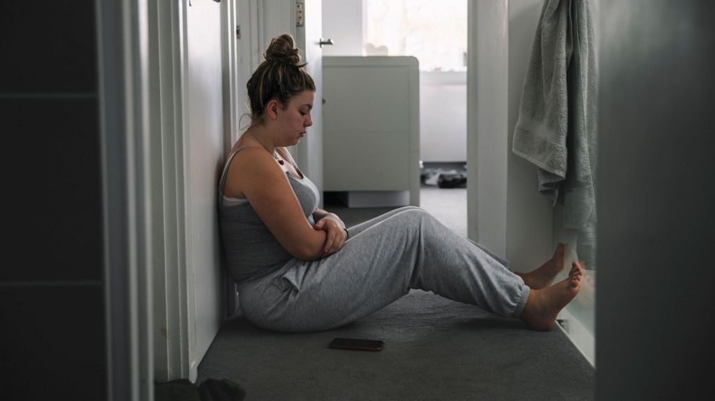 A young woman sits in the hallway of her home looking sad and lonely